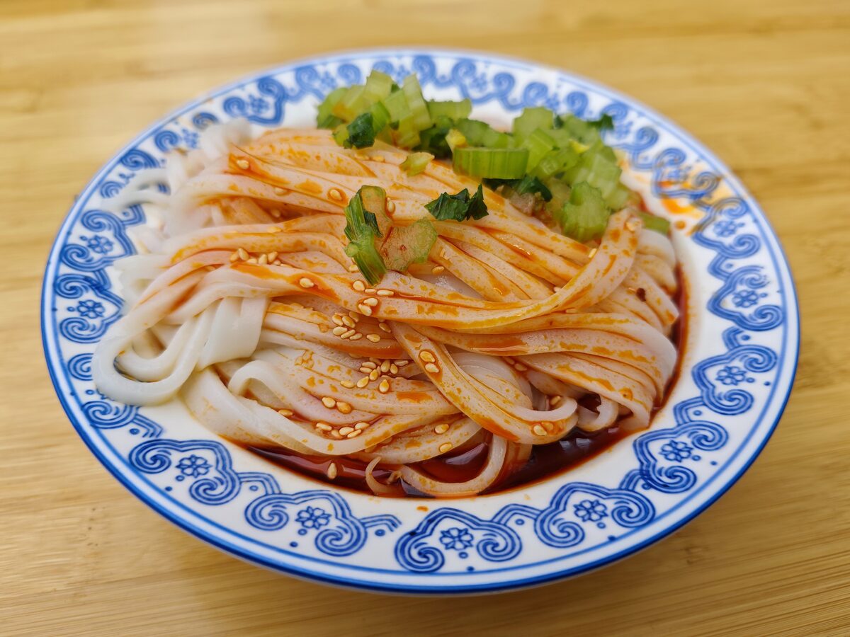 A blue-and-white plate of liangpi, wide cold noodles dressed with chili oil, sesame seeds and scallion