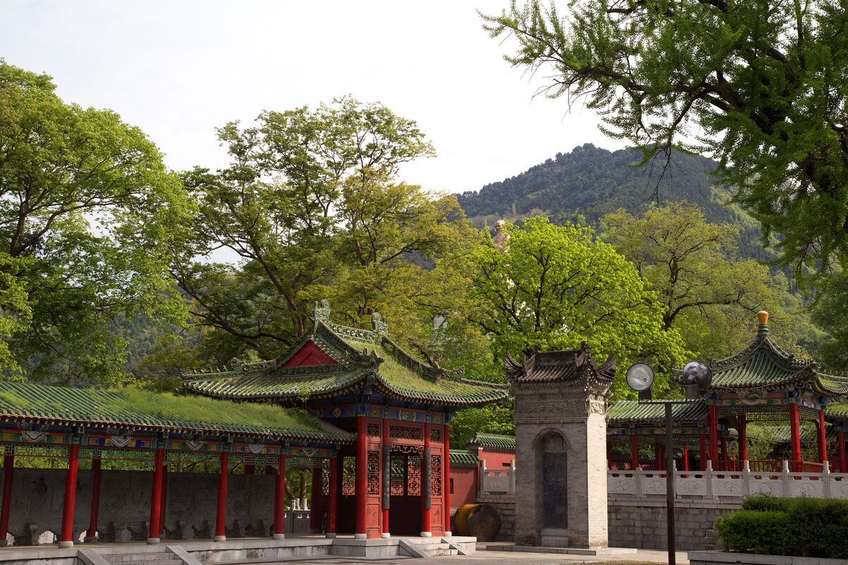 Red-pillared pavilions and a stone stele at Louguantai Taoist temple, with a wooded hill rising behind