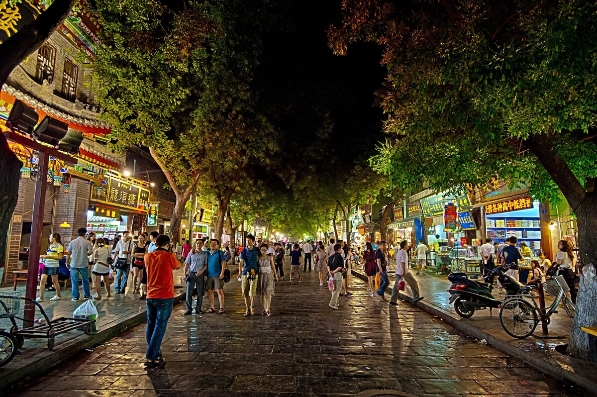 A busy street in the Xi'an Muslim Quarter at night, with shop signs glowing under the trees and crowds walking down the middle
