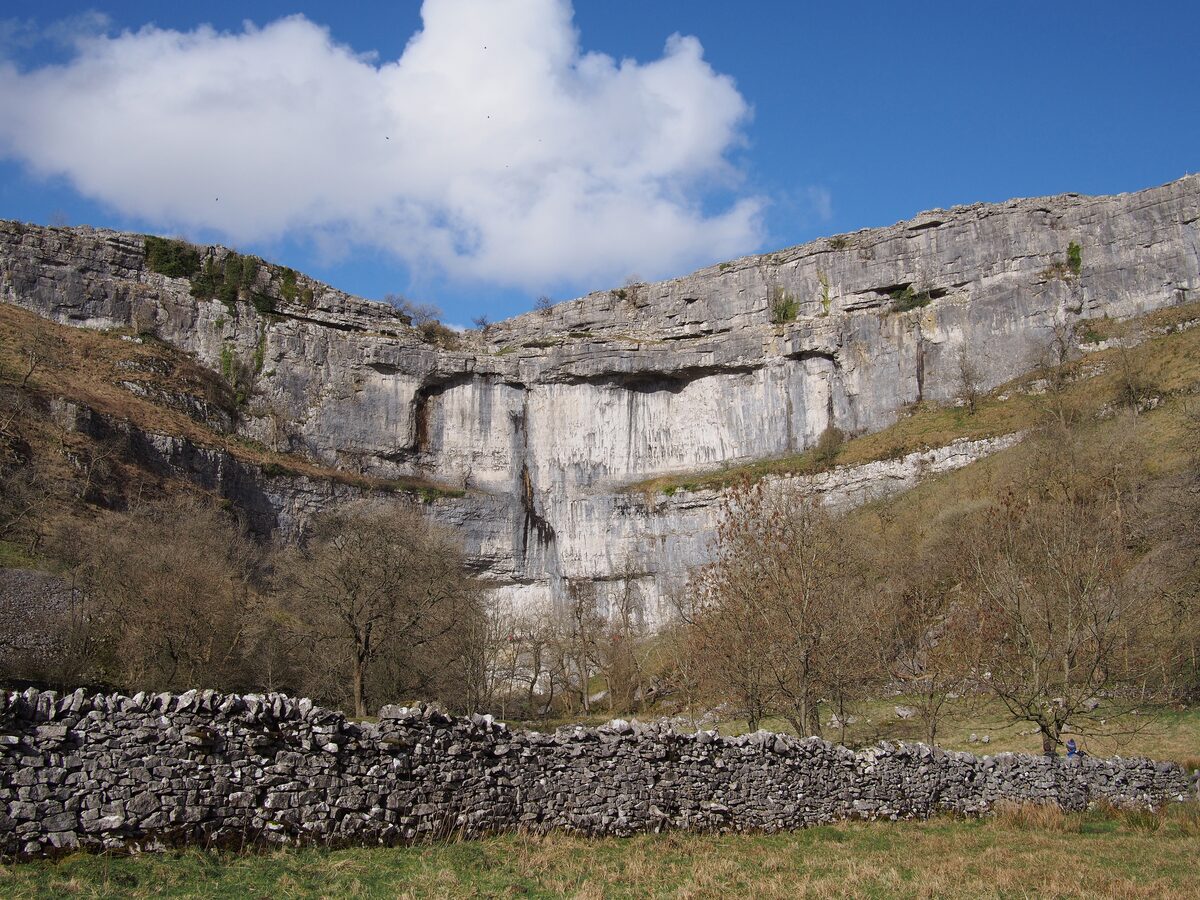 The curved limestone cliff of Malham Cove in the Yorkshire Dales, above a dry stone wall and trees