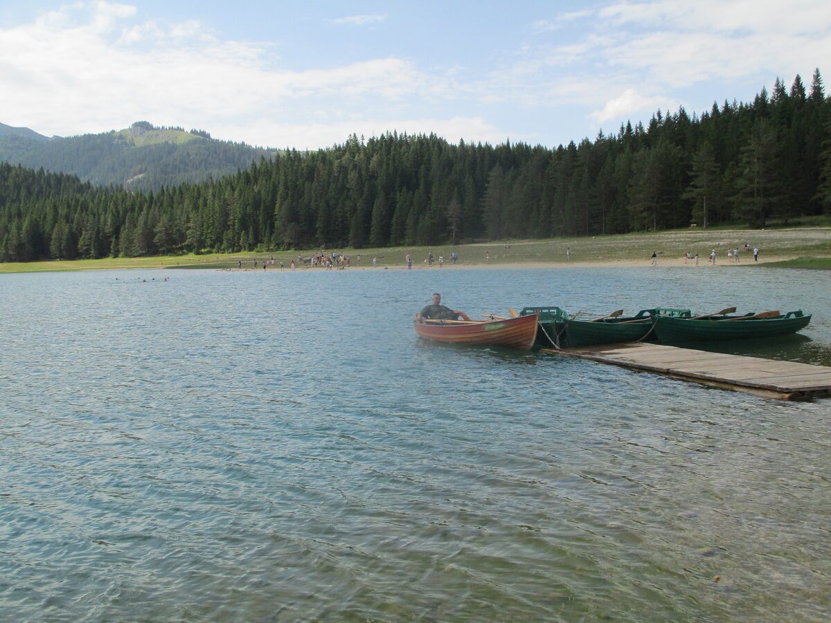 Rowing boats moored at a wooden jetty on the Black Lake, with visitors on the far shore and spruce forest behind