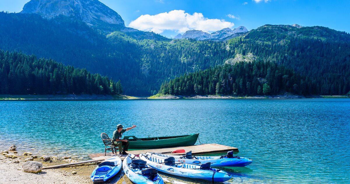 Black Lake with a green rowing boat and blue kayaks on a pebble shore, with forested slopes and a rocky peak behind the water