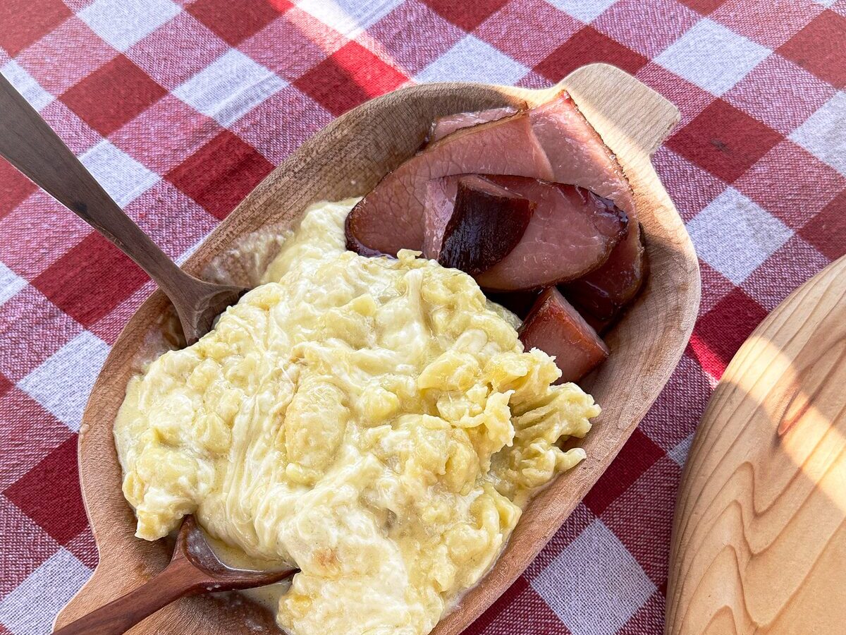 Creamy kačamak of cornmeal and cheese in a wooden bowl with slices of smoked ham, on a red and white checked cloth