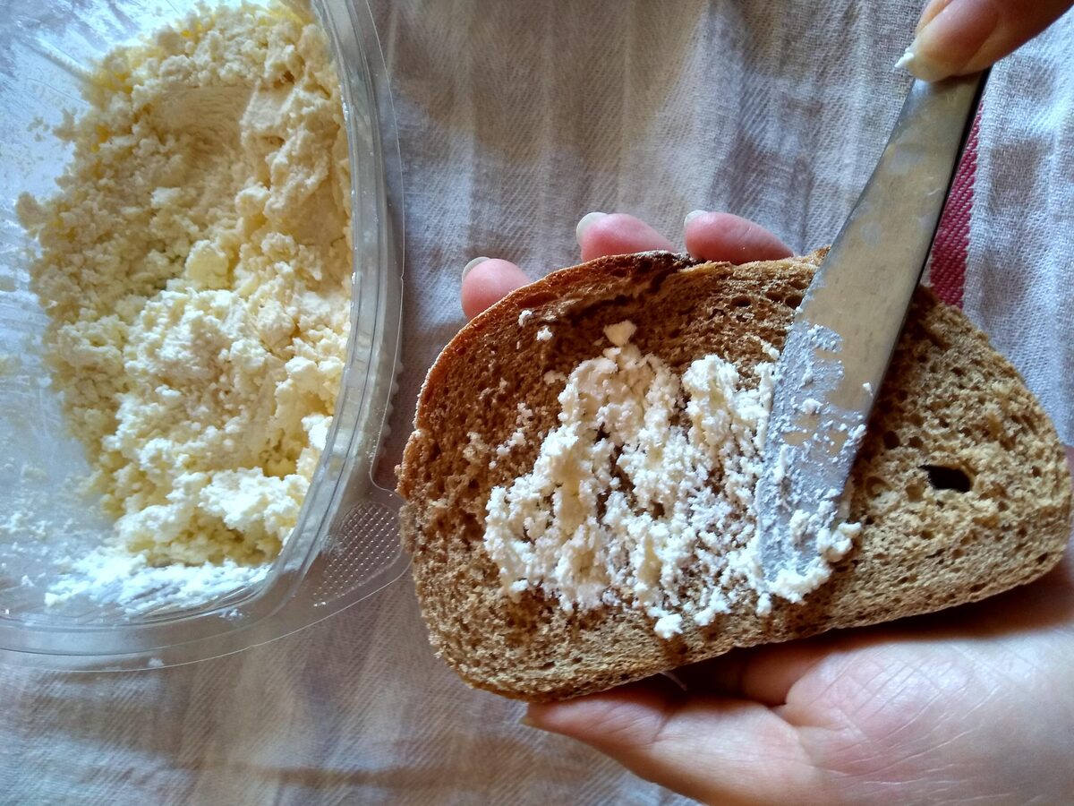 Kajmak spread thickly on a slice of dark bread held in a hand, with a tub of the creamy cheese beside it