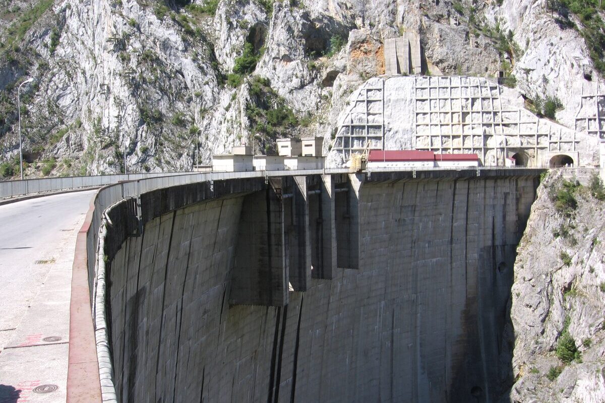 The curved concrete wall of the Mratinje dam seen from the road across its crest, with the white rock of the Piva canyon above