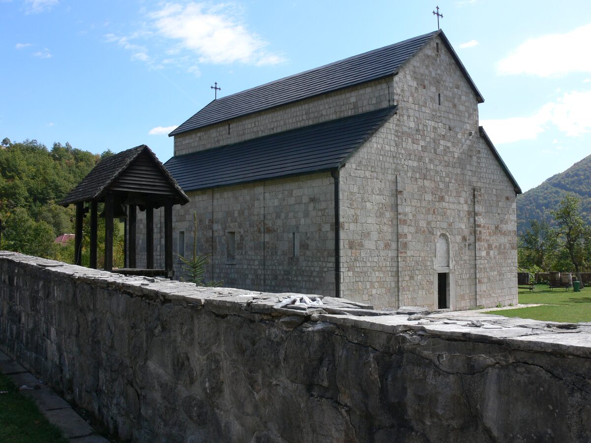 The Piva Monastery, a stone church with a dark tiled roof, a wooden canopy and a stone wall in front