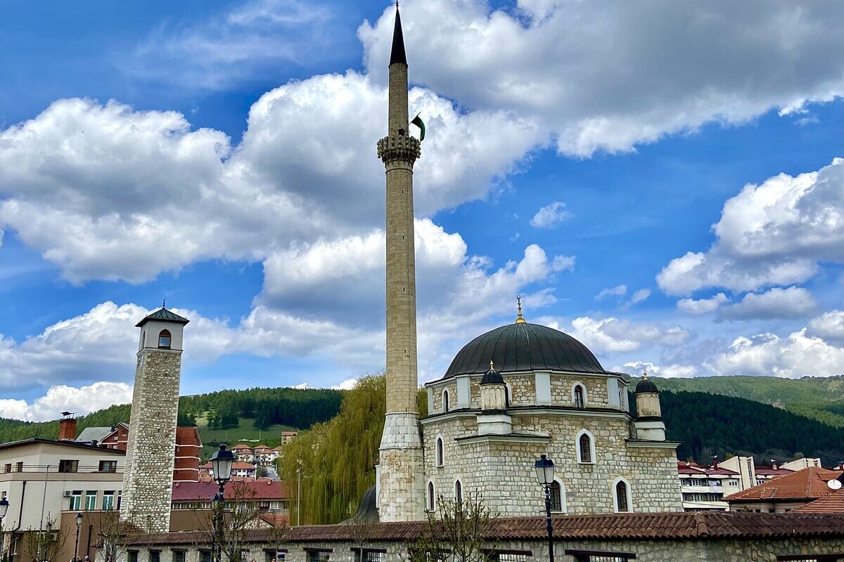 Husein-paša's Mosque in Pljevlja with its tall stone minaret and grey dome, the clock tower to the left and forested hills behind, under a blue sky with white clouds