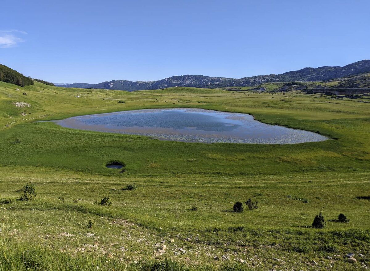 Pošćensko Lake, a shallow lake in a green meadow below the grey limestone ridges of Durmitor