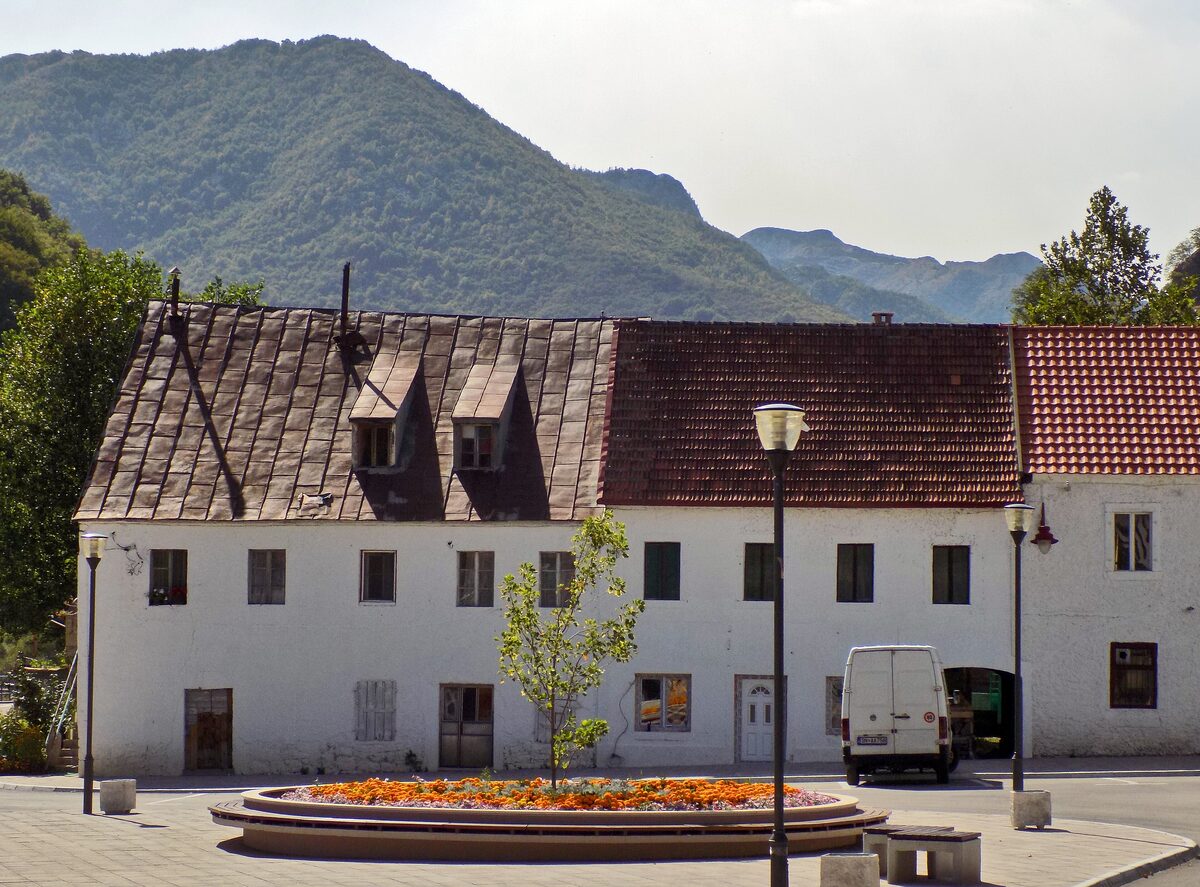 A white two-storey building with a red tiled roof on the square in the centre of Šavnik, with forested mountains behind