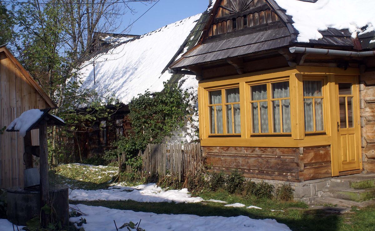 A wooden house in Chochołów with snow on its roof, a well and patches of snow in the garden
