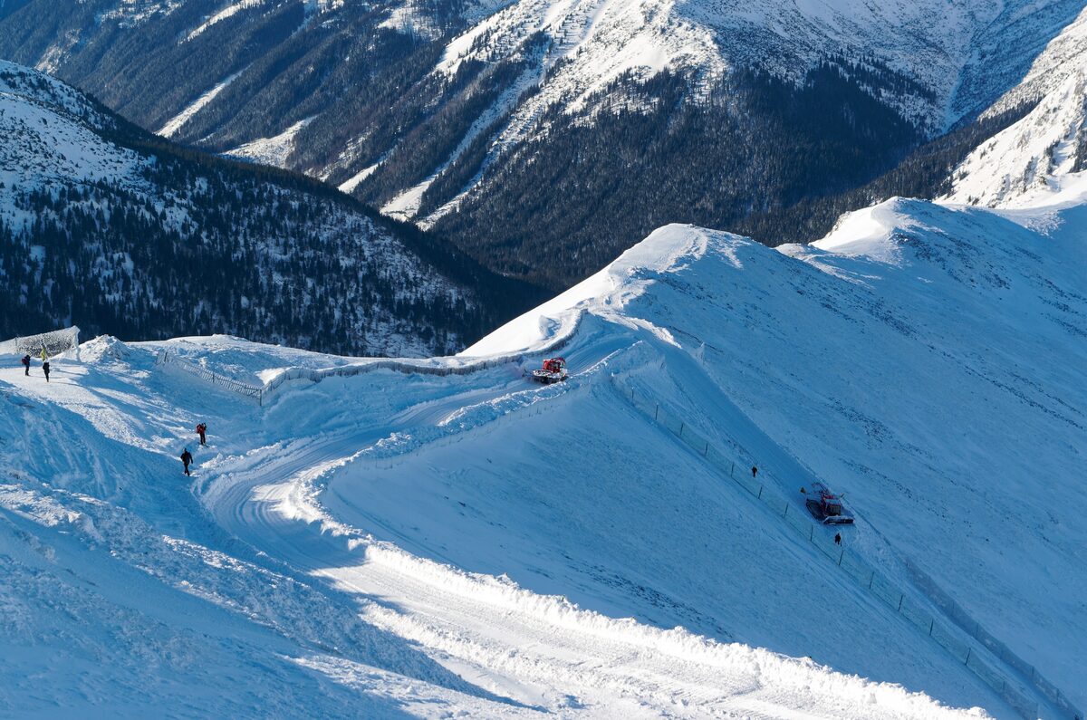A groomed snow track curling along a sunlit ridge below the top of Kasprowy Wierch, with two snow groomers and a few walkers and skiers above a forested Tatra valley