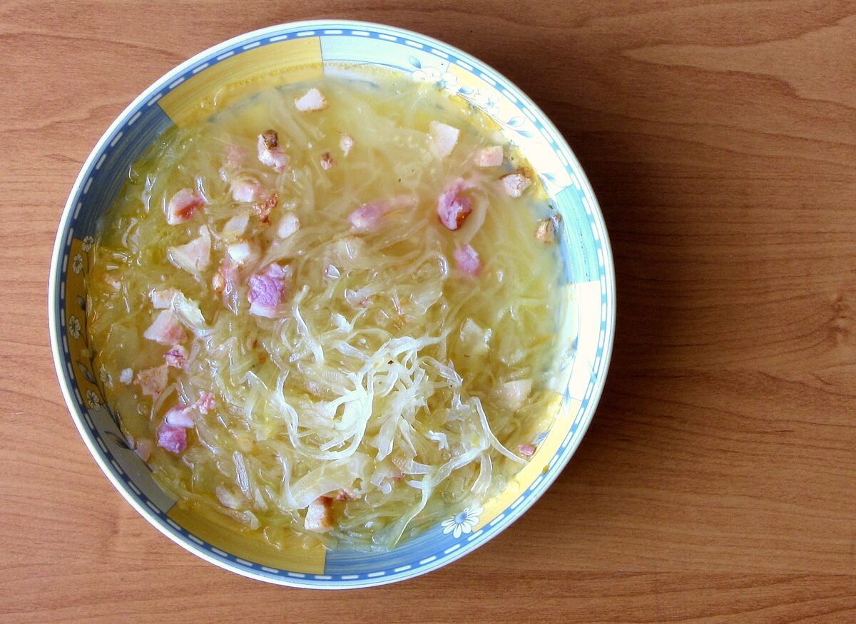 A bowl of kwaśnica, a pale sour cabbage soup with bits of meat, seen from above on a wooden table