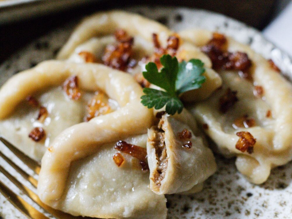 Plated pierogi topped with fried bacon bits and parsley, one dumpling cut open to show the filling