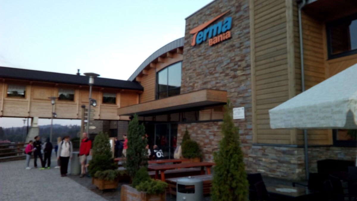 The entrance of the Terma Bania thermal complex on a summer evening, a stone and timber building with the Terma Bania sign and visitors outside