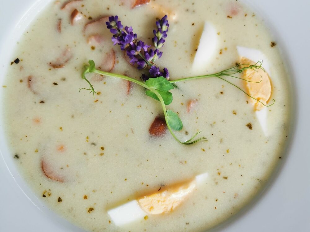 A white bowl of creamy żurek soup with halved boiled egg, sausage slices and a sprig of herbs
