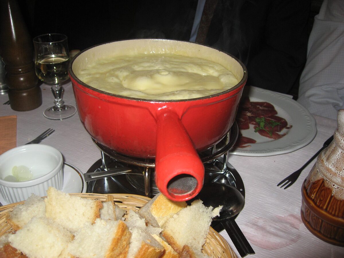 A red fondue pot of melted cheese on a burner, with a basket of bread cubes beside it