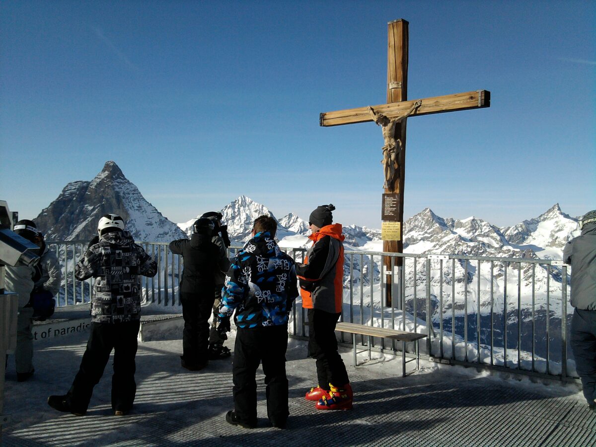 Visitors on the summit platform of Klein Matterhorn beside a wooden cross, with the Matterhorn and snowy peaks behind