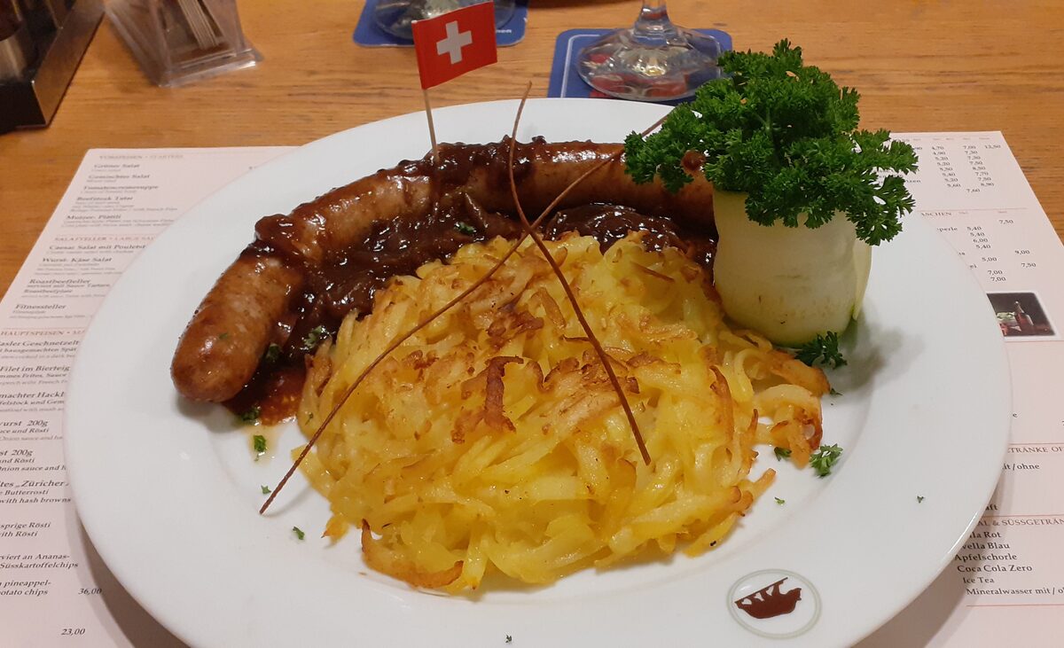 A golden rösti with a pork sausage and a small Swiss flag on a white plate