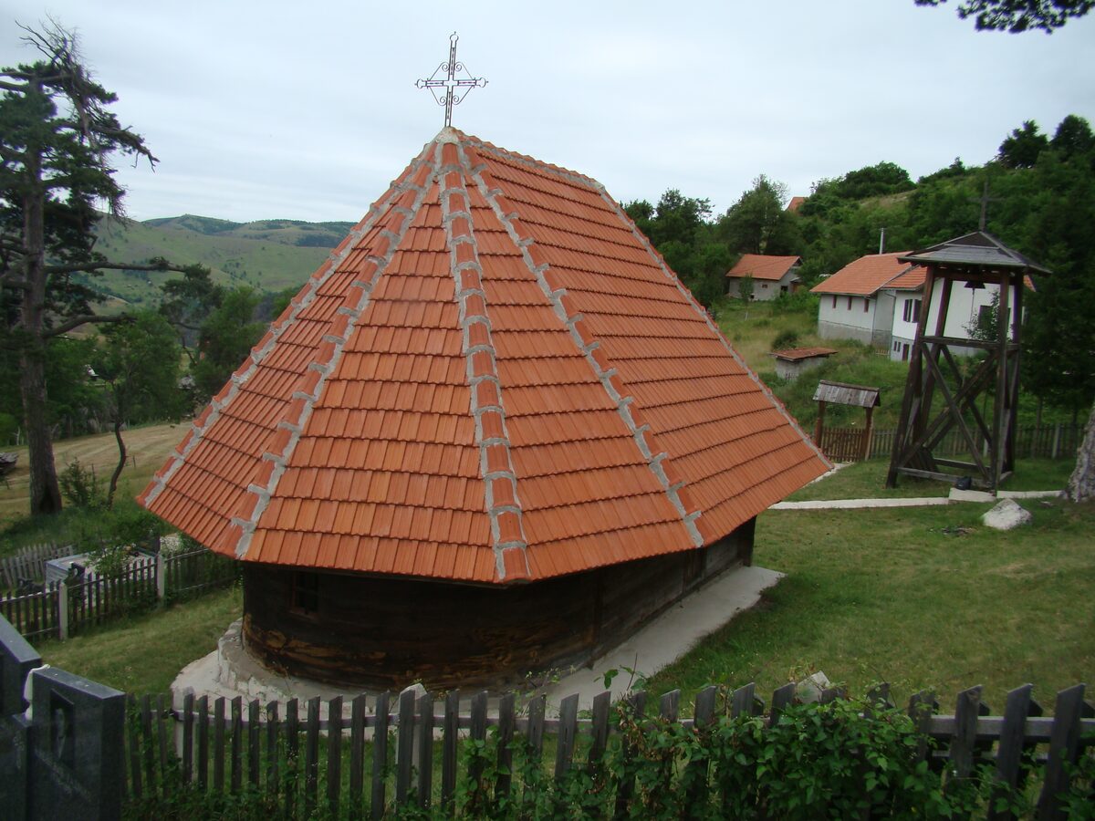 The log church in Dobroselica under a tall, steep roof of red tiles, with a wooden bell tower and village houses on the green hillside behind.