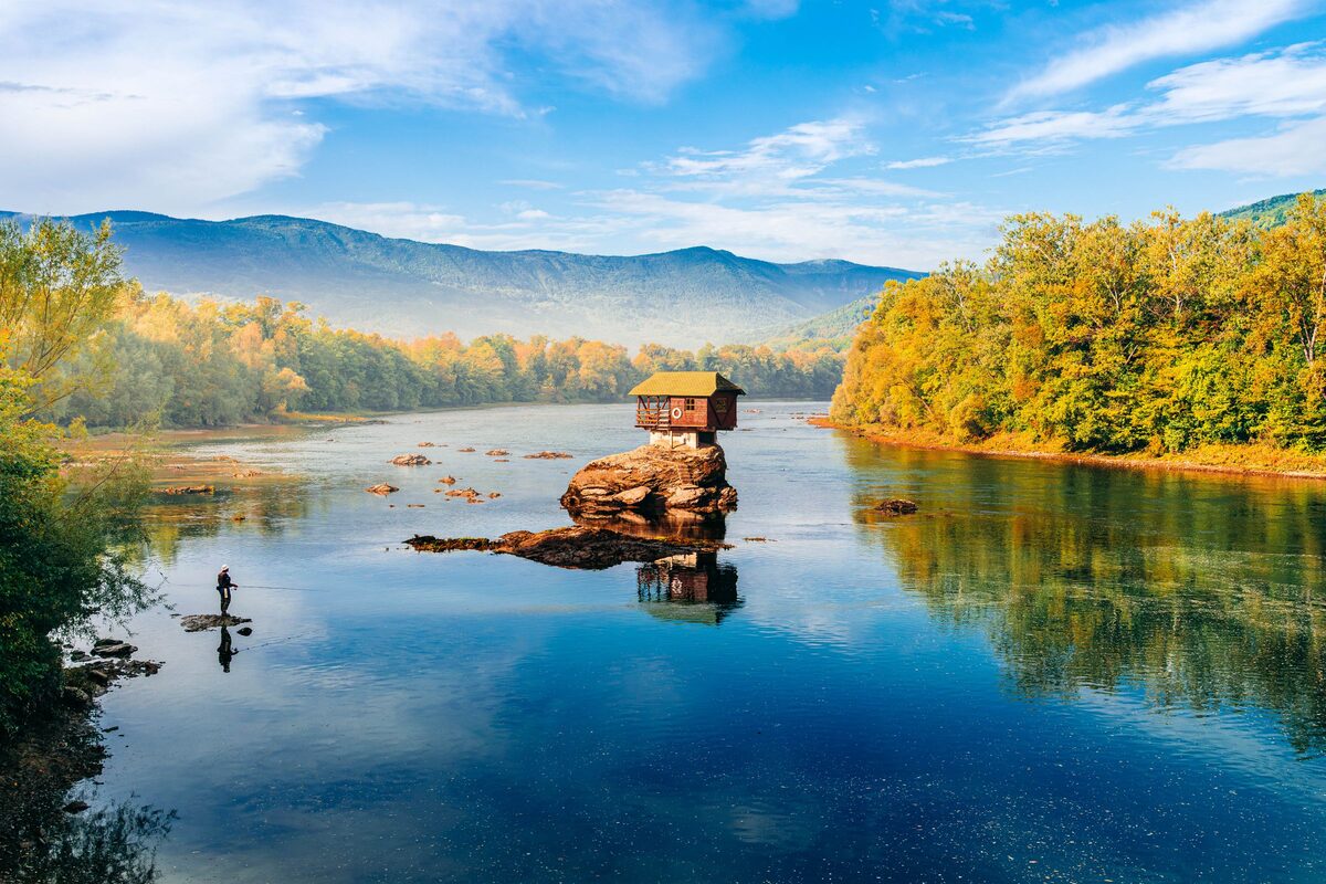 The little wooden house on a rock in the middle of the Drina, the calm river reflecting autumn trees, blue mountains and the sky.