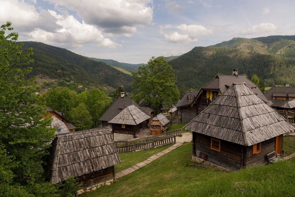 Log houses with steep shingled roofs among green lawns and trees in Drvengrad, with the wooded hills of Mokra Gora behind.