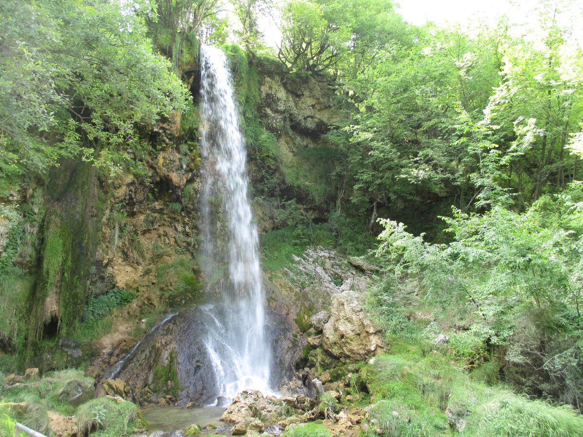 The Gostilje waterfall dropping in a single white stream over a mossy rock wall into the green forest below.