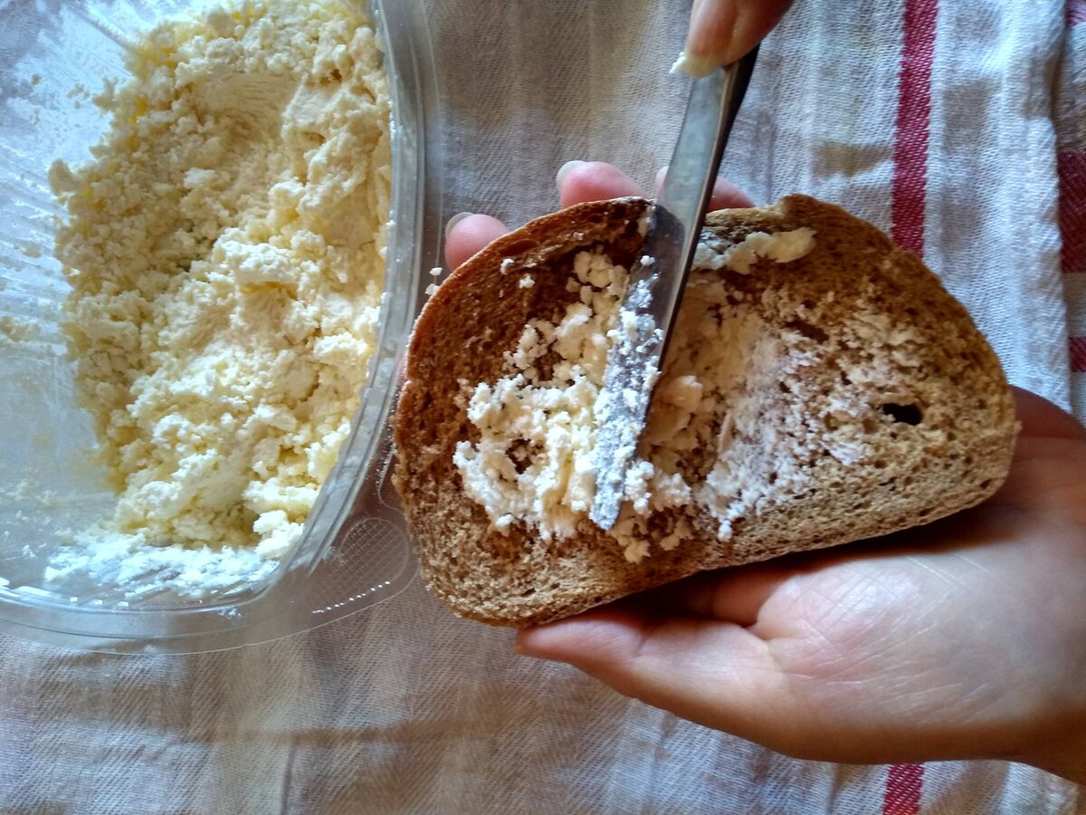 A knife spreading crumbly white kajmak onto a slice of dark bread, with a tub of kajmak beside it.