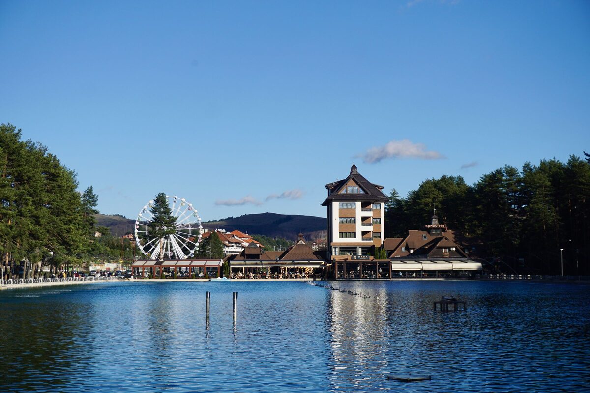 The lake in Zlatibor town on a clear day, with the Ferris wheel, a tall hotel and pine trees reflected in the blue water.