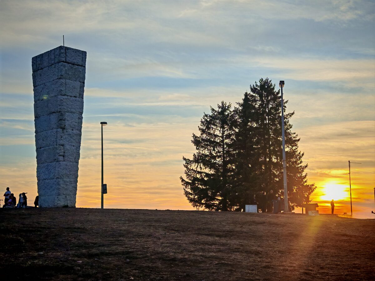 The tall white stone obelisk on top of Šumatno brdo at sunset, with two pines silhouetted against the orange sky beside it.