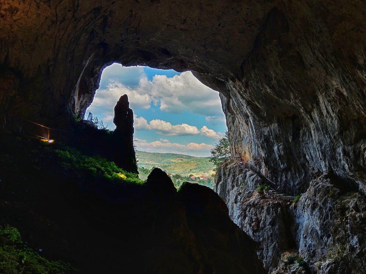 The view out through the huge arched mouth of Potpeć cave, its dark rock walls framing blue sky, clouds and green hills.