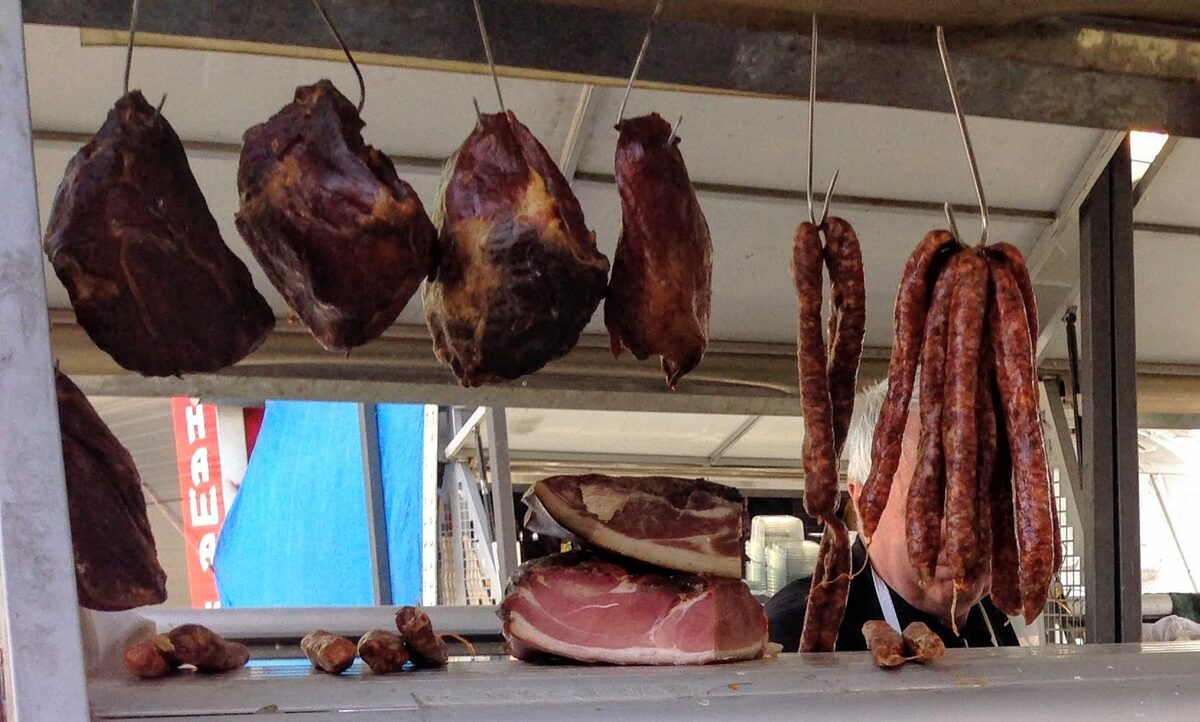 Dark pieces of dried pršut and strings of sausages hanging from hooks at a market stall, with cut pršut on the counter below.