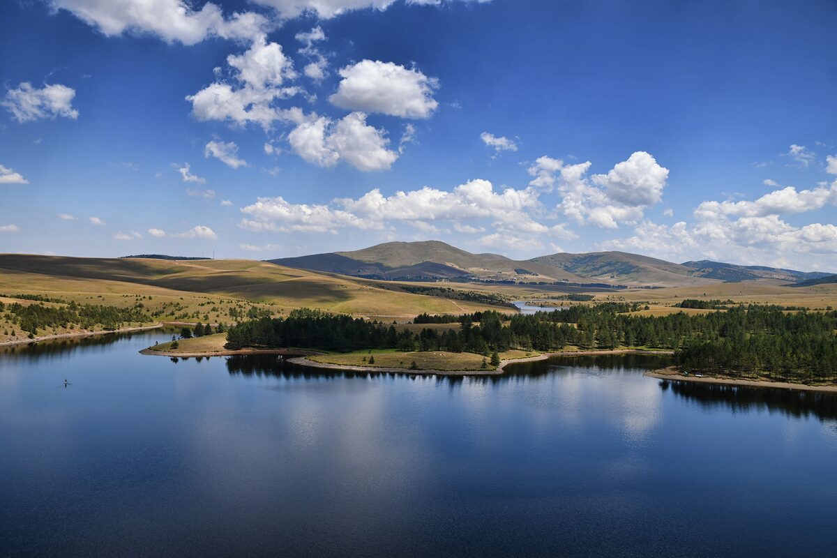 Ribnica lake mirroring white clouds and a blue sky, with pine groves on its shores and the golden meadows and hills of Zlatibor beyond.