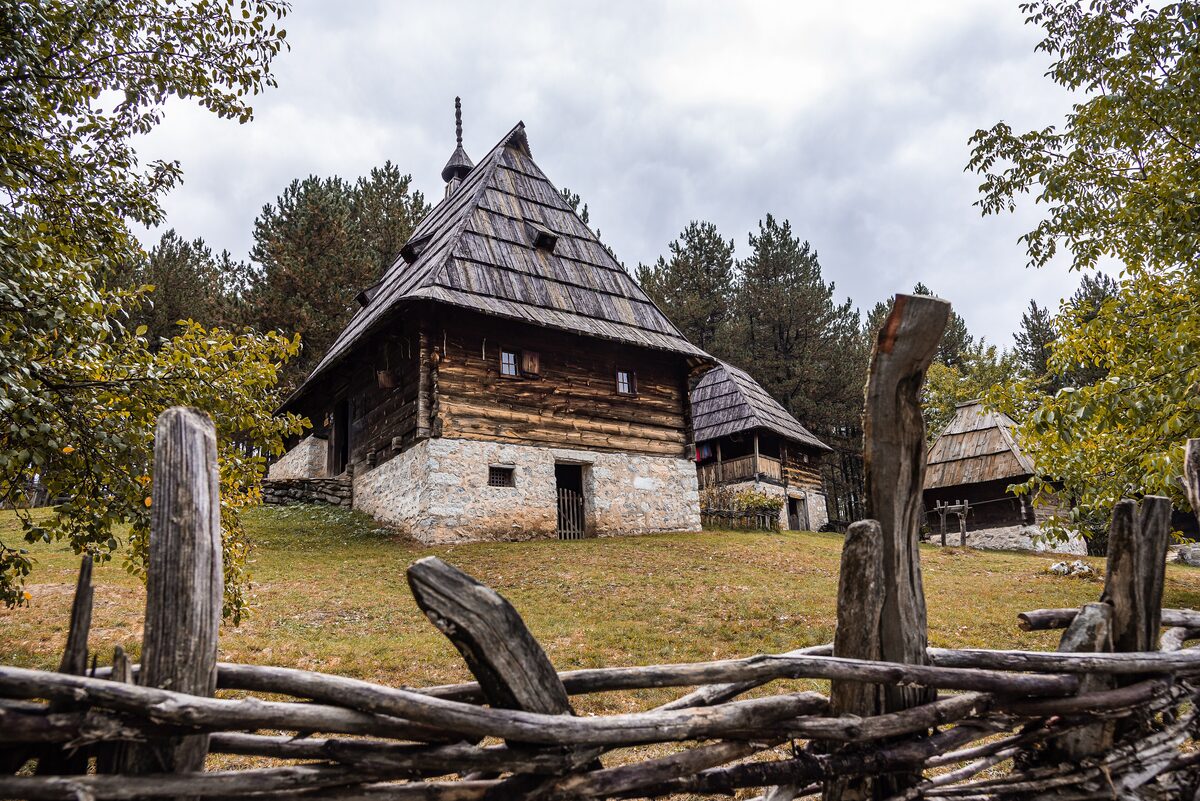 A log house with a tall shingled roof on a white stone base at the Sirogojno open-air museum, behind a fence of rough split wood.