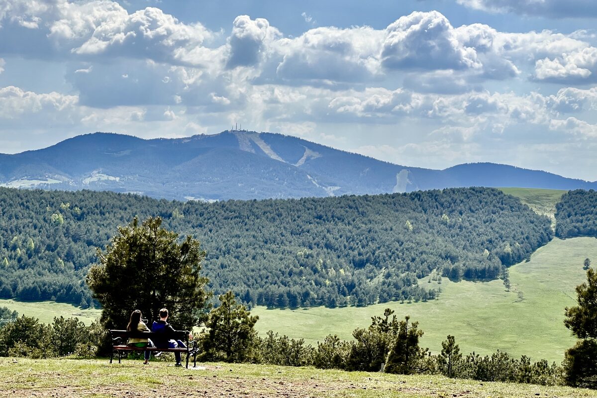 Tornik peak with its antennas and ski runs rising beyond pine forests and green meadows, two people on a bench looking at the view.