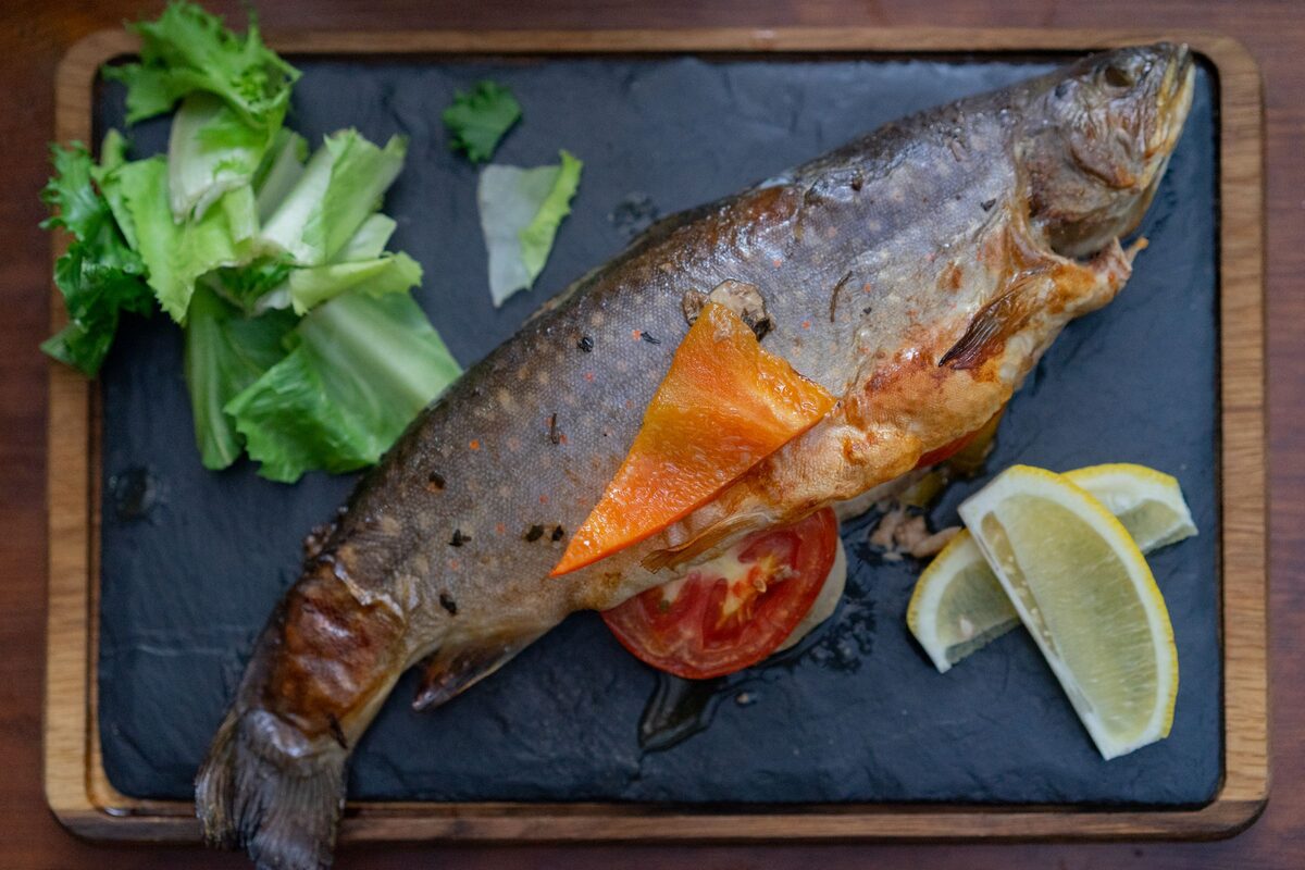 A whole grilled trout on a slate board with lemon wedges, tomato, a slice of orange and fresh lettuce.