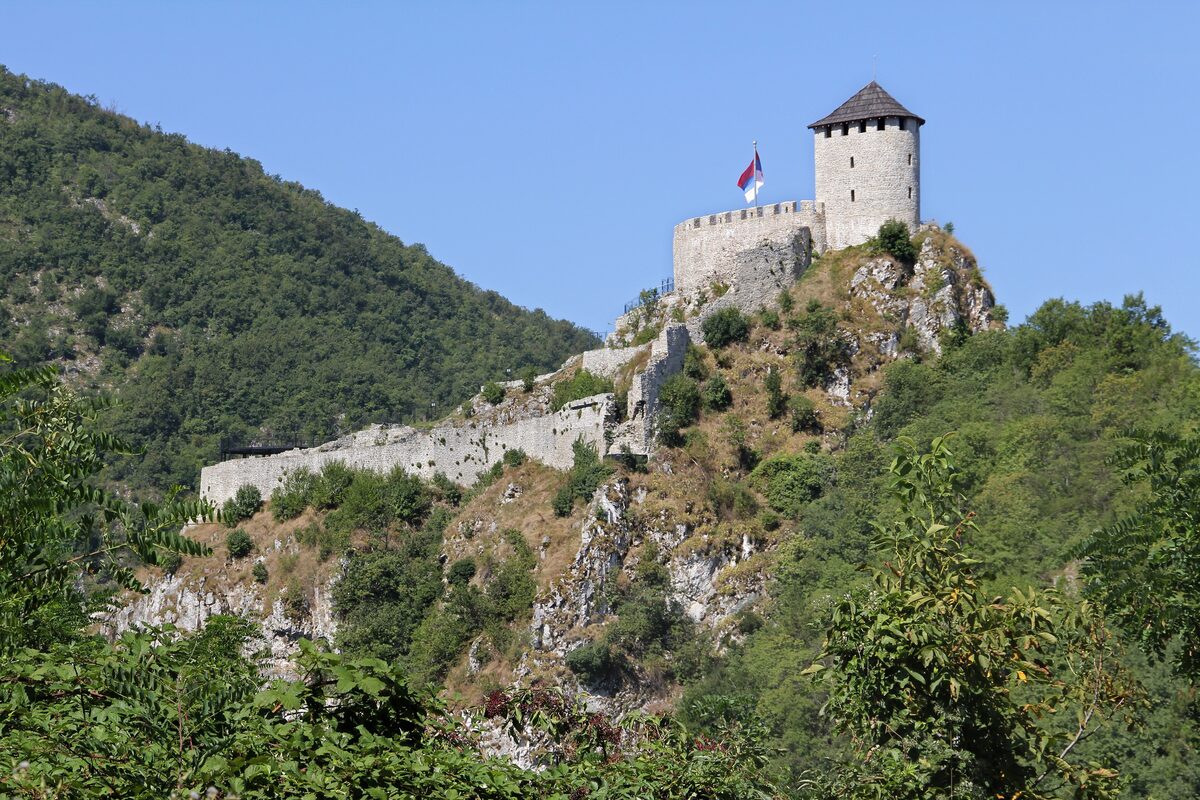 The white walls and square tower of the Užice fortress, flying the Serbian flag, on a rocky wooded crag under a clear blue sky.