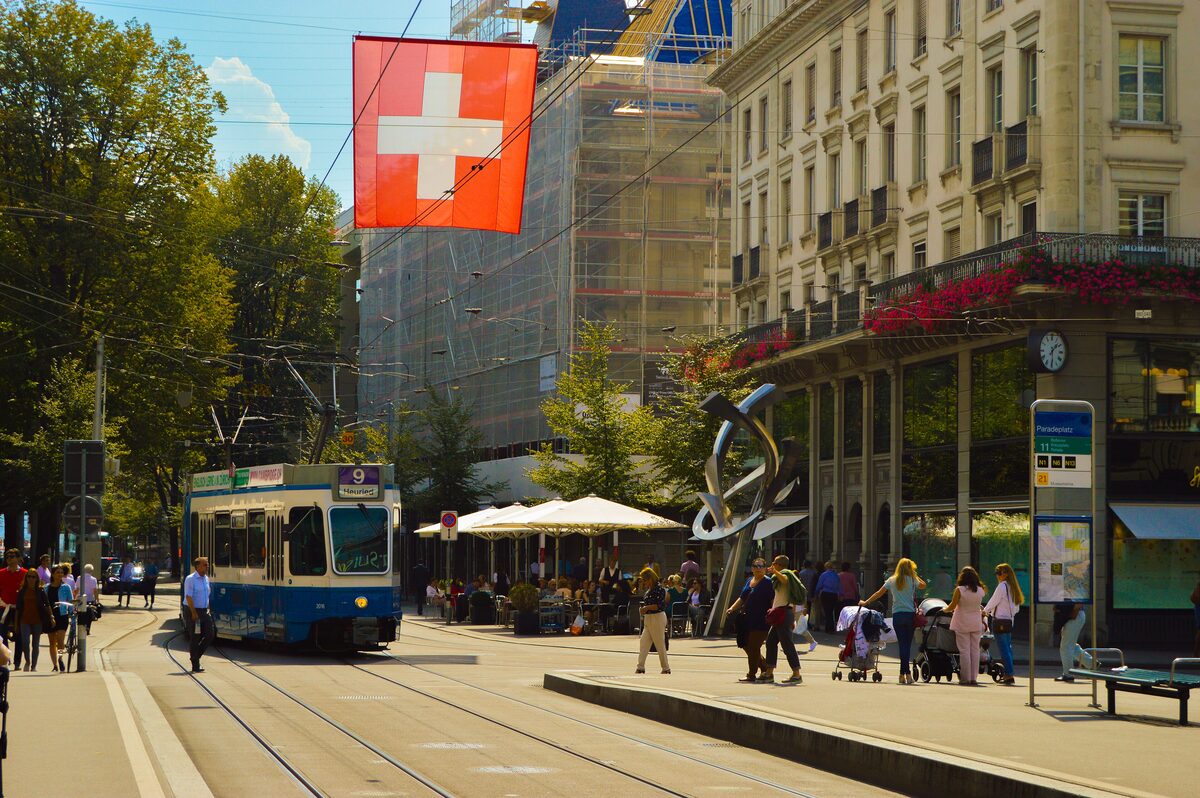 A tram on the Bahnhofstrasse below a Swiss flag, with café tables and people on the pavement