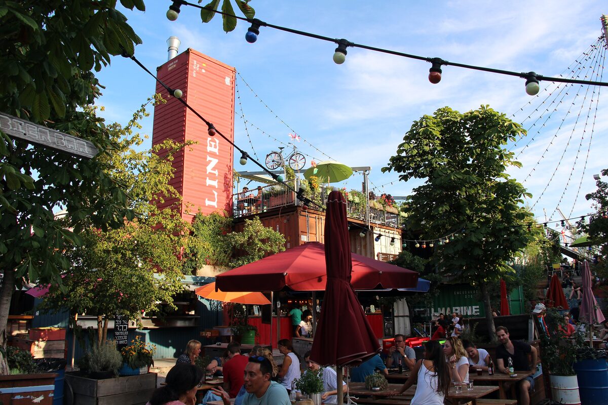 Diners at red parasols and tables under strings of lights in Frau Gerolds Garten, with a red container tower behind