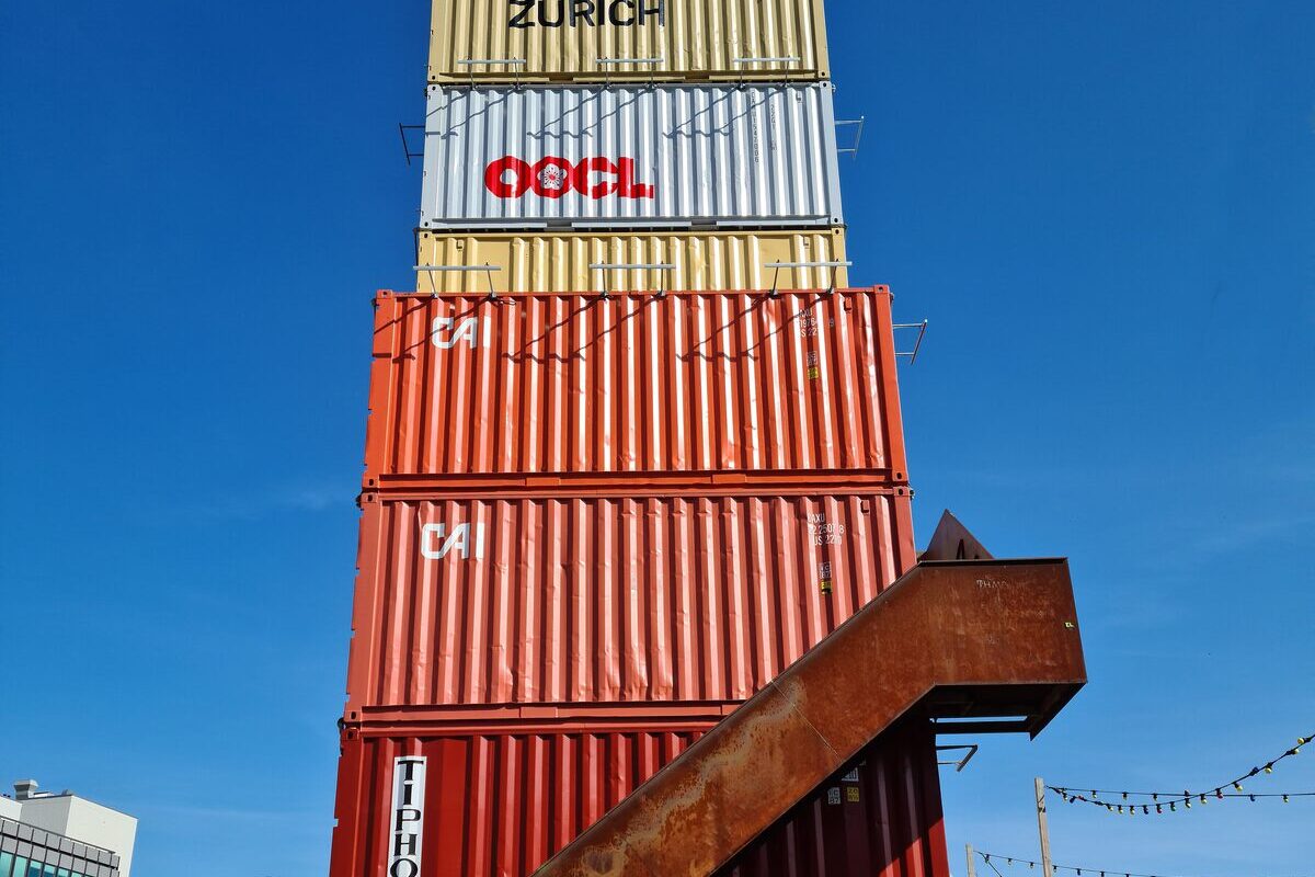 The stacked shipping-container tower of Freitag in Zurich West, red, white and sand-colored containers with a rusty steel staircase under a blue sky and a string of lights at the edge