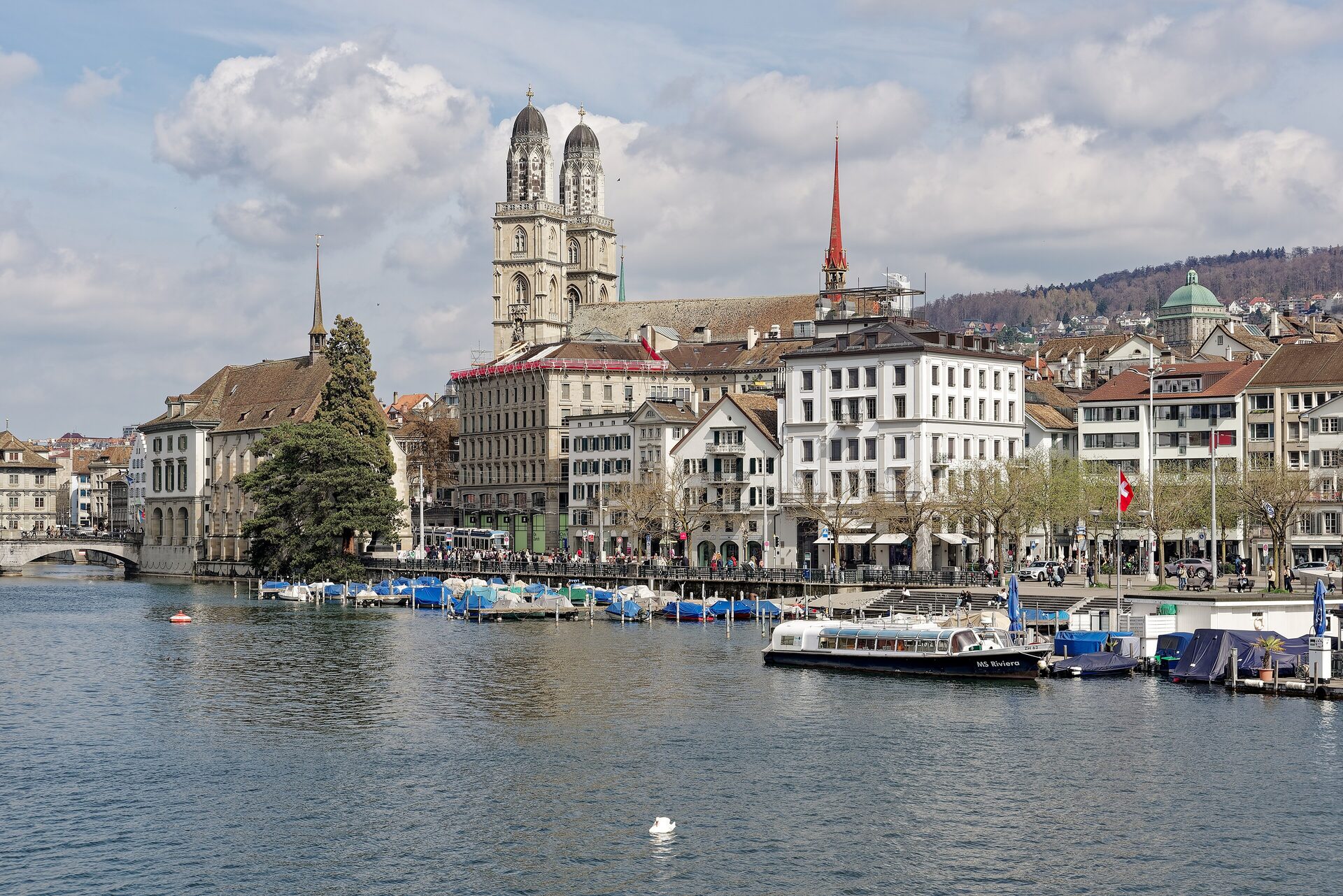 The Grossmünster towers and the old town above the Limmat, seen across the river from the lake end, with moored boats and a river boat in front