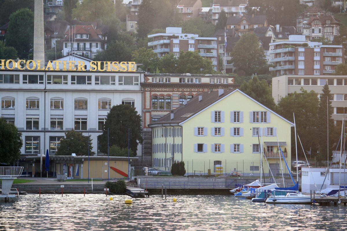 The old Lindt and Sprüngli factory buildings with the Chocolatier Suisse lettering on the Kilchberg shore, seen from Lake Zurich with sailboats at the jetty; the Home of Chocolate museum is not in the frame
