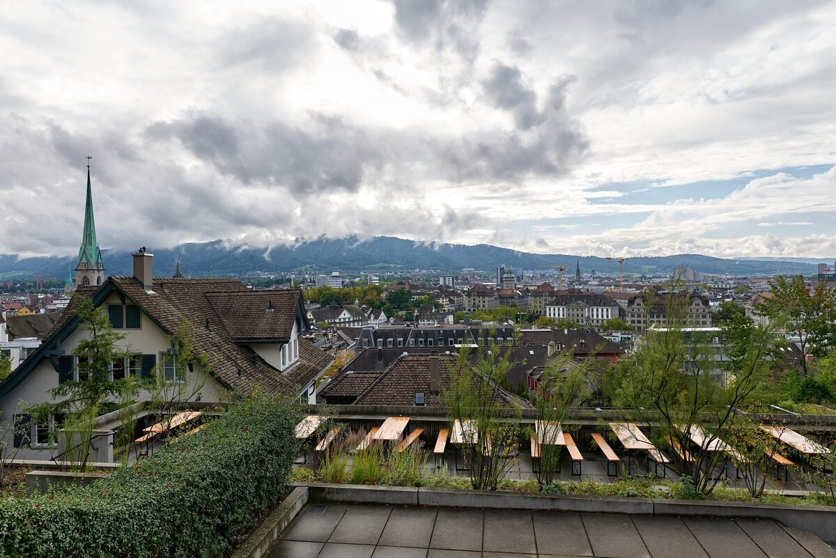 The view from the Polyterrasse over the old town roofs and a green church spire, with picnic tables on the terrace below and the wooded Üetliberg ridge under heavy clouds
