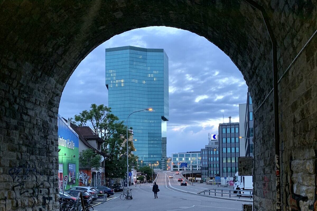 The blue-green glass Prime Tower seen through a stone railway arch at dusk, with a street and offices below
