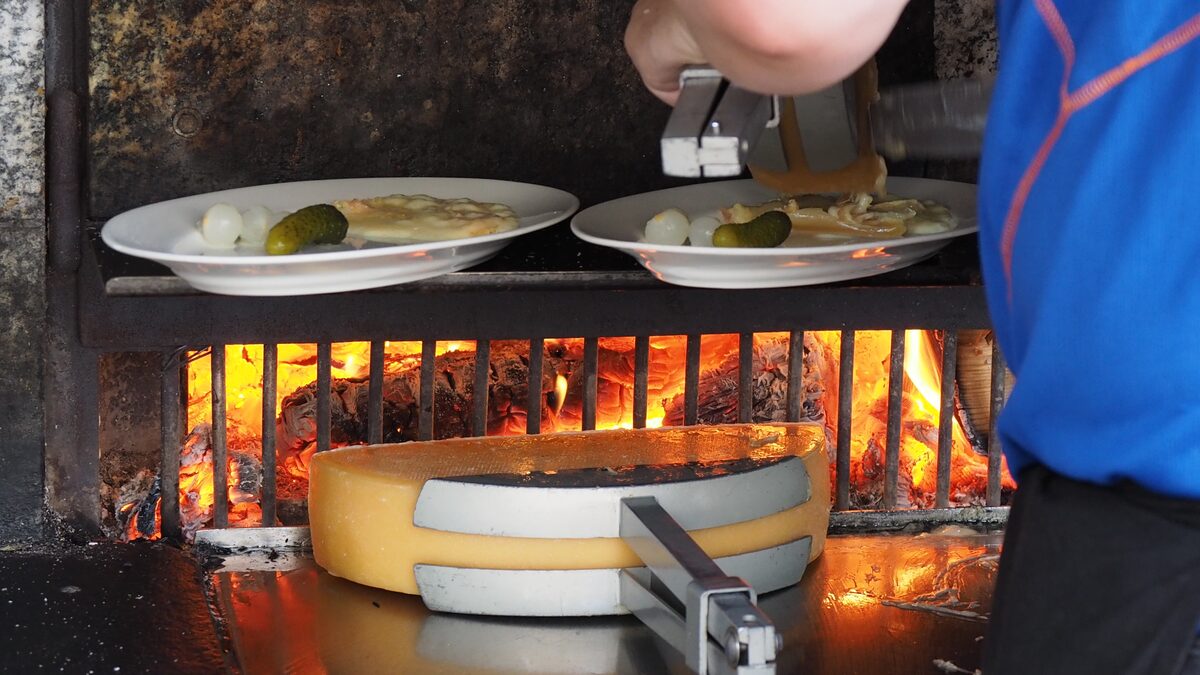 Half a raclette wheel melting in front of an open fire, with a cook scraping cheese onto plates of potatoes and pickles