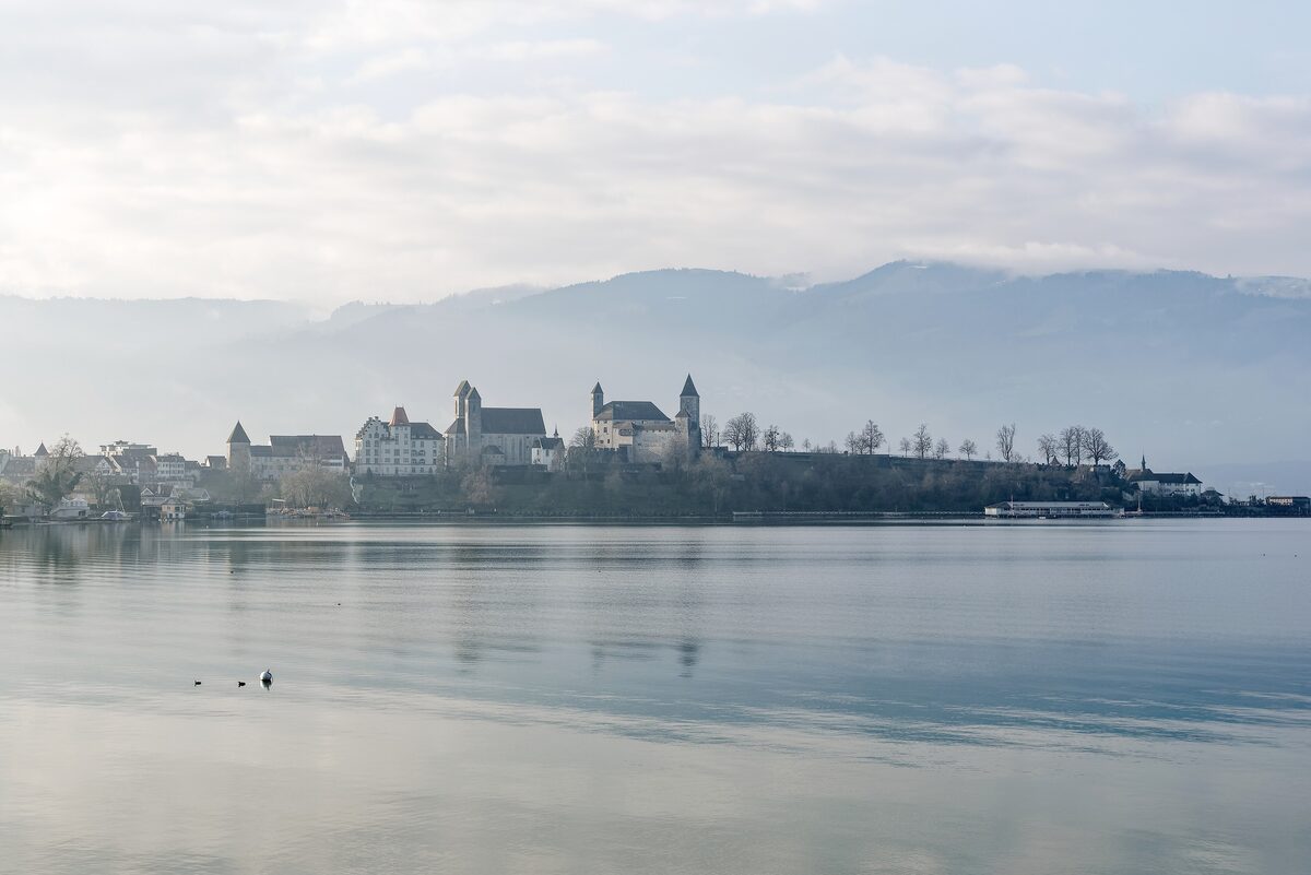 Rapperswil's castle and church towers on the far lakeshore in soft winter haze, with the hills of the Obersee behind