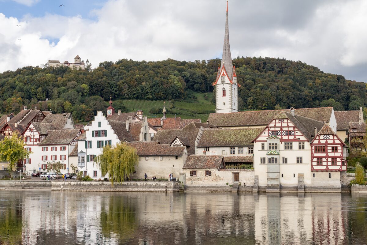 The painted old town of Stein am Rhein on the river, with a tall church spire and a castle on the wooded hill