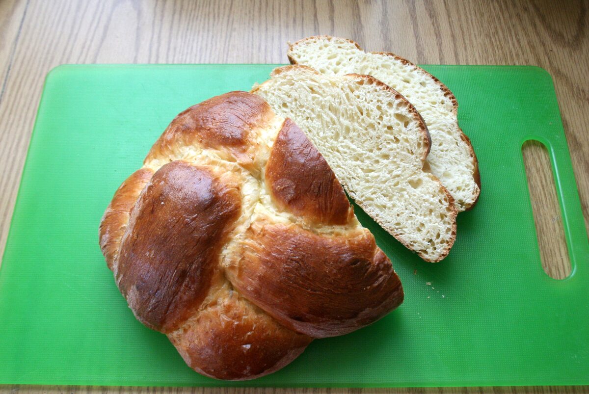 A braided Zopf loaf on a green board, one slice cut to show the soft white crumb