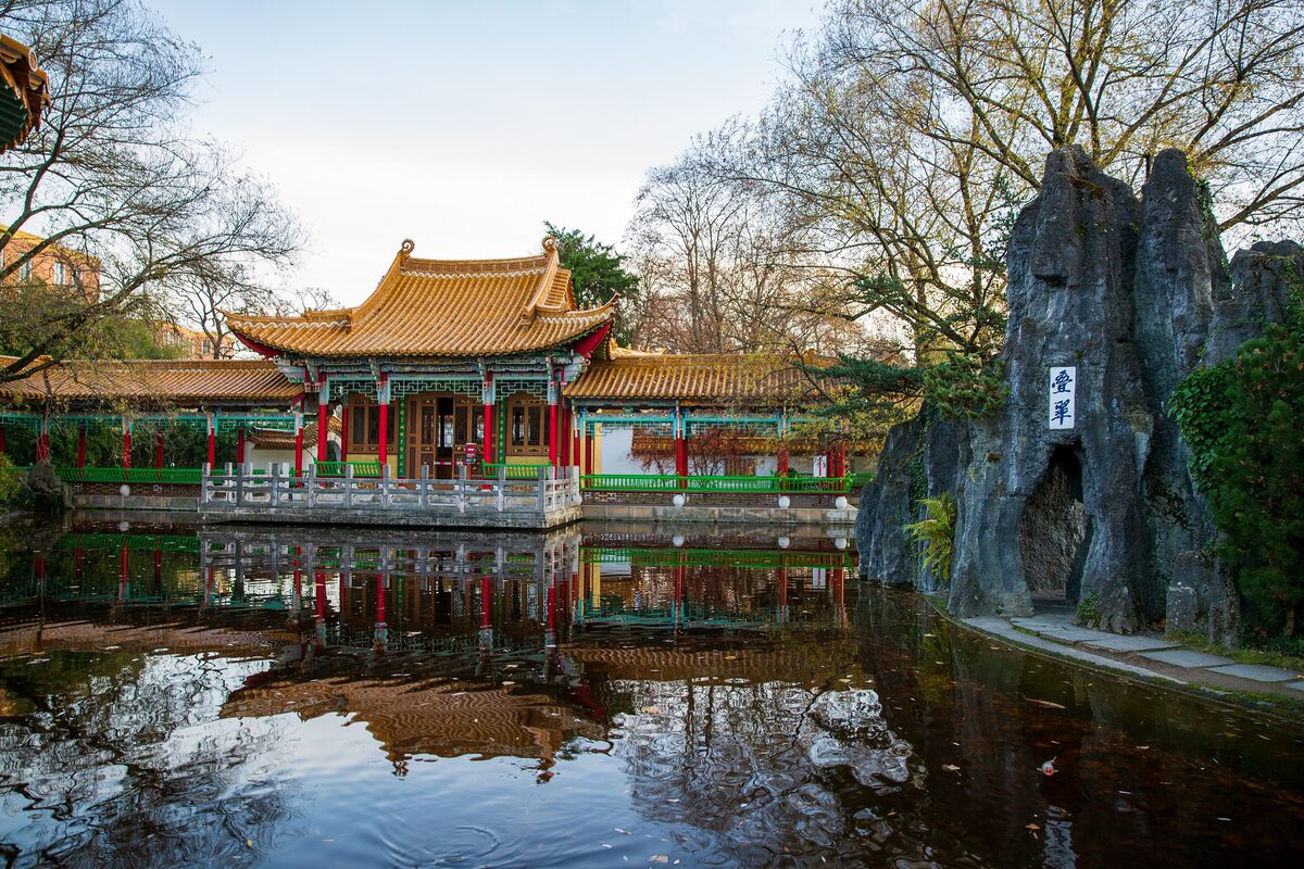 A Chinese pavilion with a yellow tiled roof reflected in the pond of the Chinese Garden at the Zürichhorn, with bare trees around it